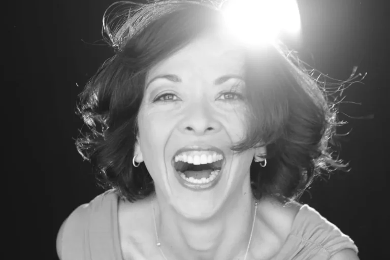A woman with shoulder-length hair smiles widely at the camera; bright light shines behind her head, creating a dramatic effect.
