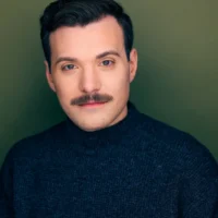 A man with dark hair and a mustache wears a dark sweater and poses in front of a plain olive green background.