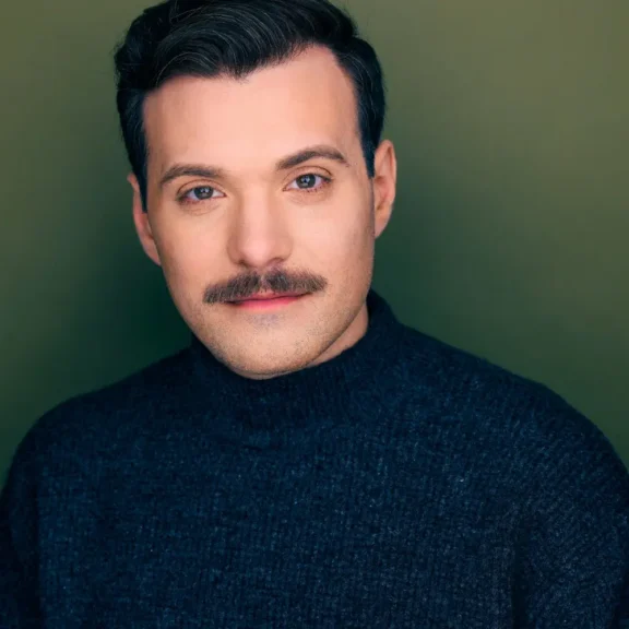 A man with dark hair and a mustache wears a dark sweater and poses in front of a plain olive green background.