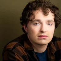 A person with curly brown hair, wearing a brown and black plaid shirt over a blue top, looks at the camera with a neutral expression against an olive-green background.