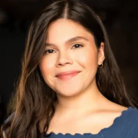 A woman with long brown hair and light skin smiles softly at the camera. She is wearing a blue top and gold hoop earrings, with a dark, out-of-focus background.