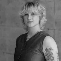 A person with short, wavy hair and a sleeveless top stands against a plain background, showing a detailed bird tattoo on their upper arm.