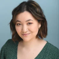 Person with short brown hair, wearing a green patterned top and a nose ring, smiles softly at the camera against a plain light blue background.