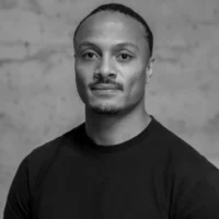 A man with short hair and a trimmed mustache and goatee wears a plain dark T-shirt, posing against a plain, textured background. The image is in black and white.