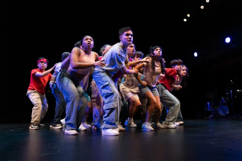 A group of young people in casual clothes stand close together on a dark stage, appearing tense and focused, under stage lighting.