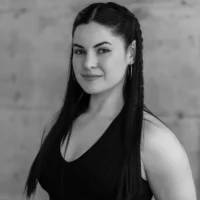A woman with long dark hair styled in braids, wearing a sleeveless top and hoop earrings, stands in front of a plain, blurred background.