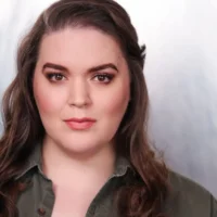 A person with long brown hair and light makeup wears a green collared shirt and looks directly at the camera against a light background.