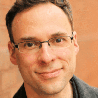 A man with short brown hair and glasses smiles slightly while standing in front of a blurred brick wall.