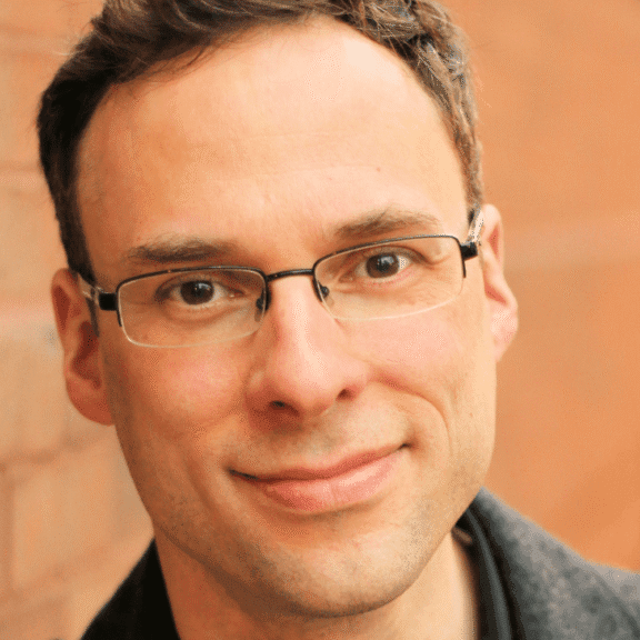 A man with short brown hair and glasses smiles slightly while standing in front of a blurred brick wall.
