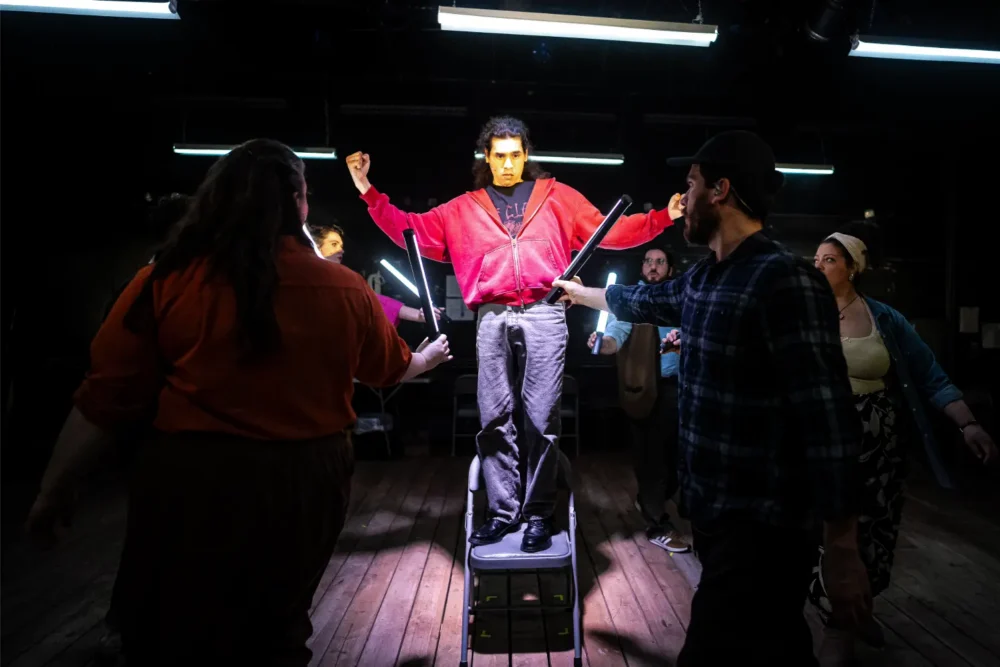 A person in a red jacket stands on a chair with arms raised, surrounded by others holding light tubes pointed toward them on a wooden stage.