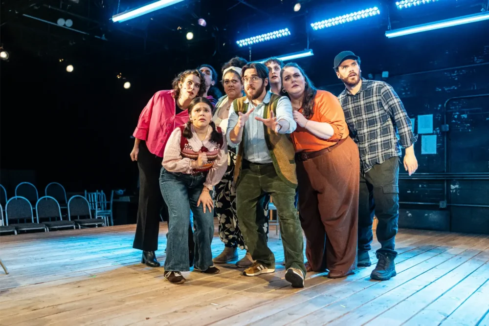 A group of actors on a wooden stage, looking up with expressions of surprise and concern.