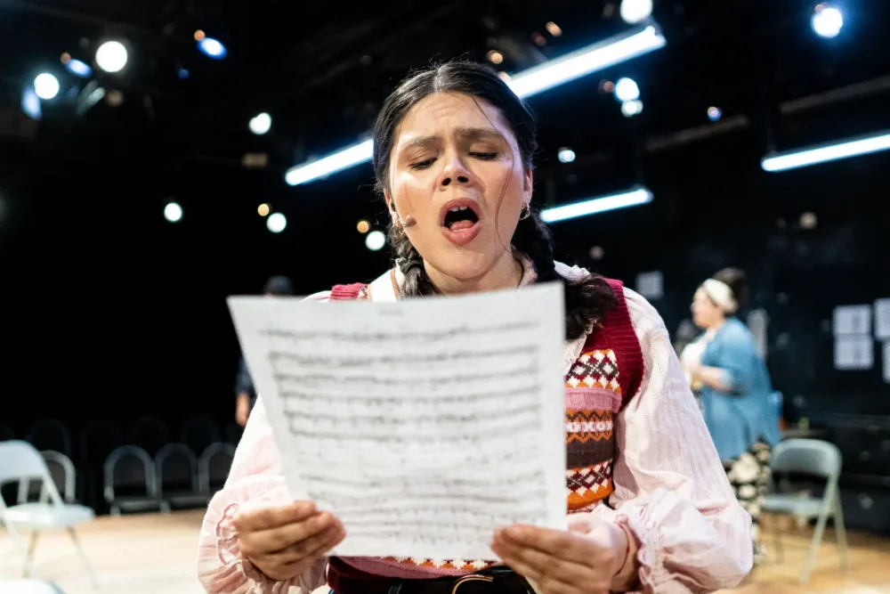 A woman with braided hair sings while holding sheet music in a brightly lit rehearsal space with empty chairs and people in the background.