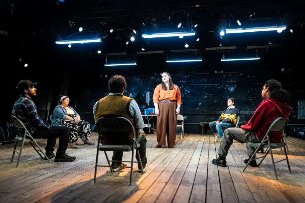 Six people are seated in a circle on a wooden stage; one person stands in the center speaking, under bright overhead lights.