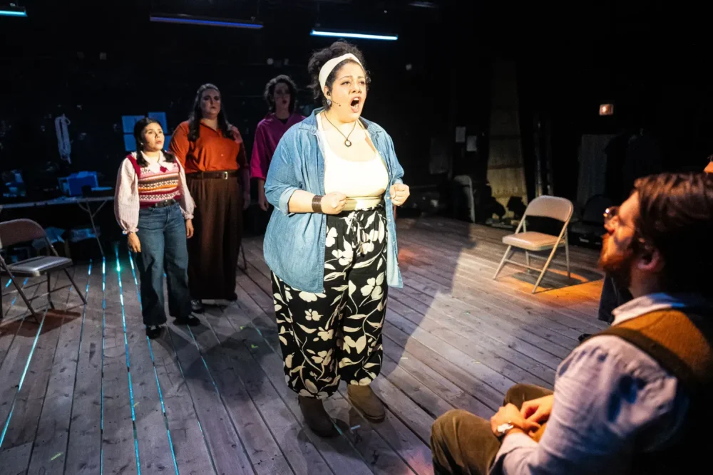A woman stands center stage singing, with three people behind her and a seated person in front, on a wooden-floored theater set with scattered chairs.
