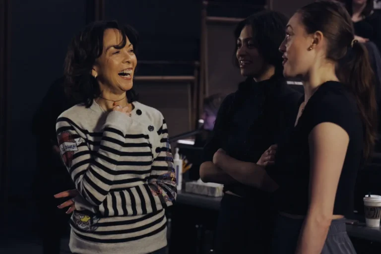 Three women stand indoors, engaged in conversation; one is laughing while the other two listen and smile.