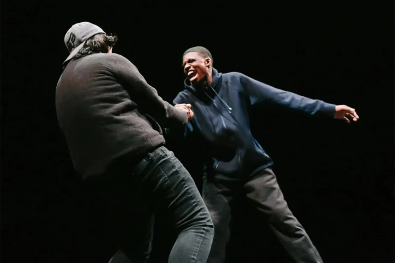 Two people are interacting on a dark stage; one is wearing a cap and sweater with their back to the camera, and the other is smiling broadly in a hoodie and pants.