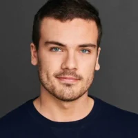 A young man with short brown hair, light beard, and blue eyes wears a navy blue shirt and small earrings, posing against a plain dark background.