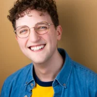 A person with short curly hair and glasses smiles at the camera, wearing a denim jacket over a yellow and navy shirt, in front of a tan background.
