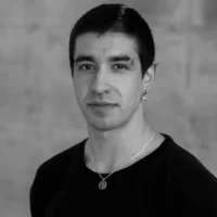 A person with short dark hair, wearing a black shirt, earrings, and a necklace, poses against a plain blurred background.
