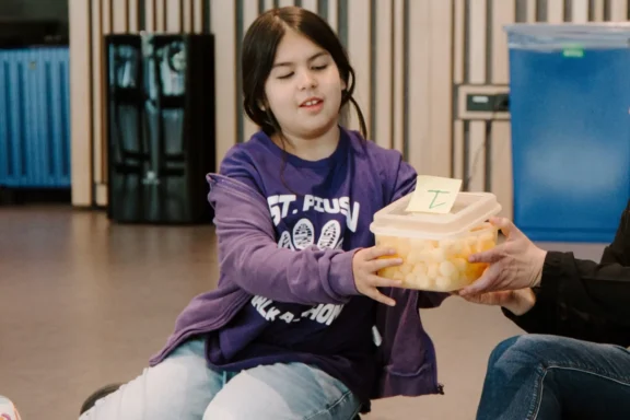 A girl in a purple shirt and jacket holds a plastic container filled with yellow balls, which has a sticky note with the letter "E" on top.