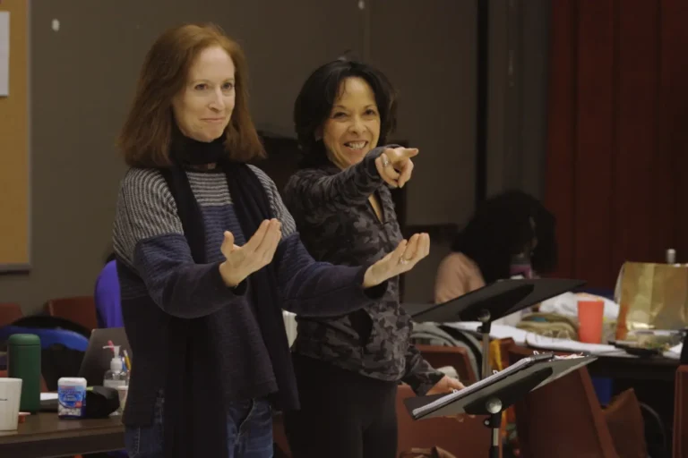 Two women stand indoors by music stands; one gestures with open hands, the other points forward and smiles. Items and people are visible in the background.