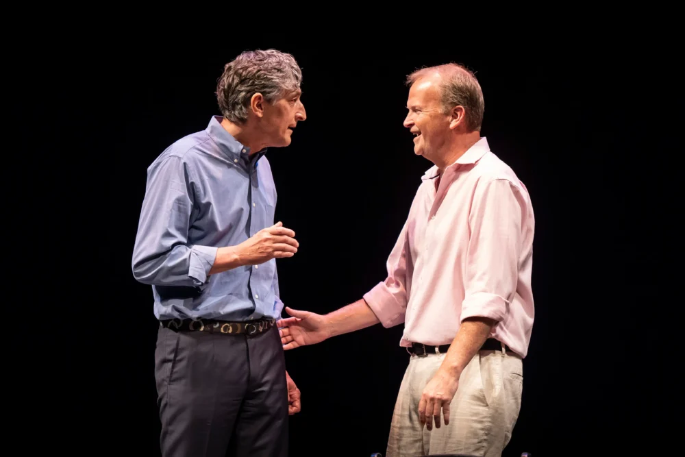 Two middle-aged men stand facing each other and talking on stage against a black background. One man gestures with his hand while the other smiles.