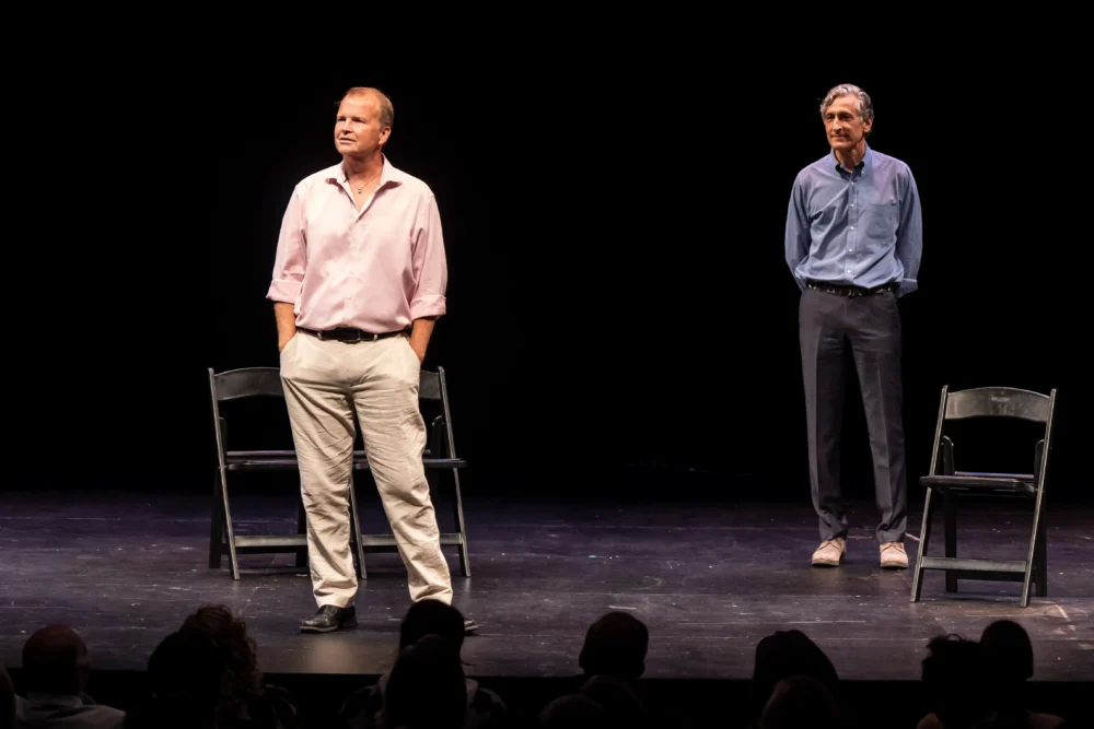 Two men stand on a stage with two chairs, one man in a light shirt and pants in the foreground, the other in a blue shirt and darker pants in the background, facing an audience.