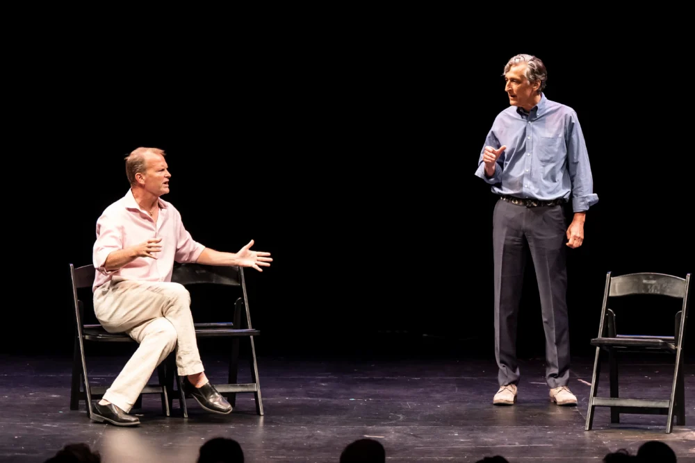 Two men perform on stage; one sits and gestures while speaking, the other stands and listens. Three empty chairs are arranged on the stage against a black background.