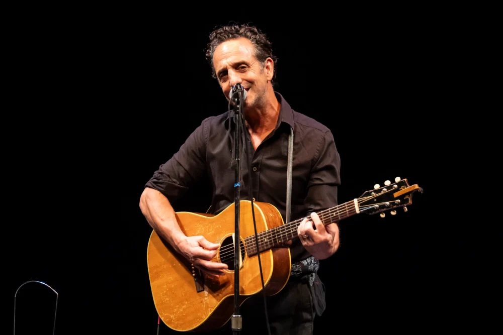 A man in a black shirt plays an acoustic guitar and sings into a microphone on stage against a black background.