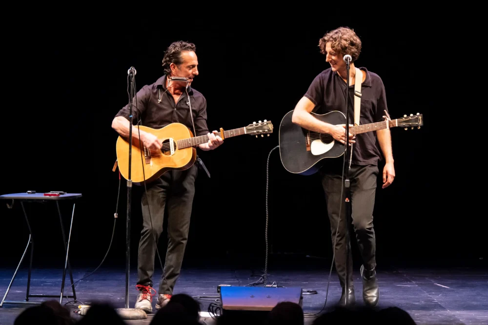 Two musicians dressed in black play acoustic guitars and stand on a stage with microphones and a small table, against a dark background.
