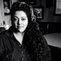 A person with long, curly hair and a streak of light color poses indoors, wearing a button-up shirt. The setting includes framed pictures on wood-paneled walls and a round table.