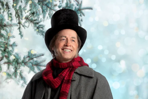 A man dressed as a nineteenth-century character, wearing a top hat and a red scarf against a snowy pine tree background.