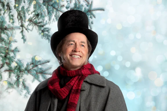 A man dressed as a nineteenth-century character, wearing a top hat and a red scarf against a snowy pine tree background.