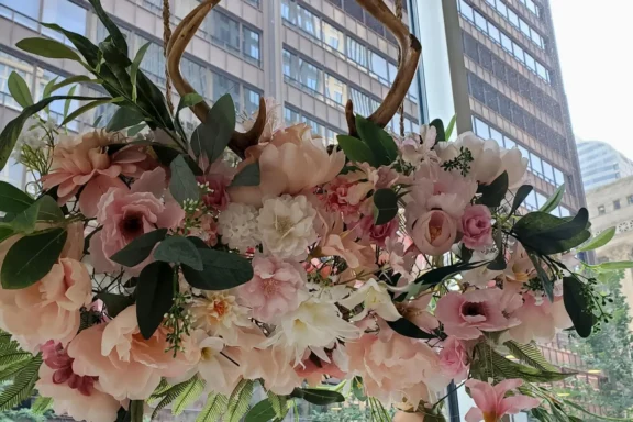 A floral arrangement of pink and white flowers with green leaves, accented by decorative antlers, displayed indoors with tall office buildings seen through the window behind.