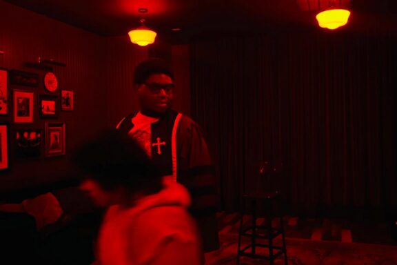 Two people in a dimly lit room with red lighting; one stands near a stool, wearing a robe with a cross, while the other moves past in the foreground.