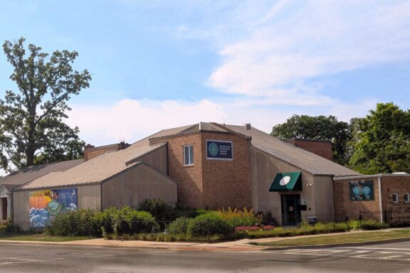 A brick building with metal roofing, multiple signs, a mural on one wall, a green awning over the door, and landscaping in front, set at a street corner under a partly cloudy sky.