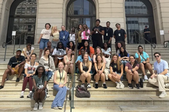 A large group of young adults sits and stands on the steps outside a stone building with arched windows, posing for a group photo.