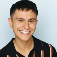 Person with short dark hair smiles at the camera, wearing a black collared shirt with brown and white vertical stripes. Light blue background.