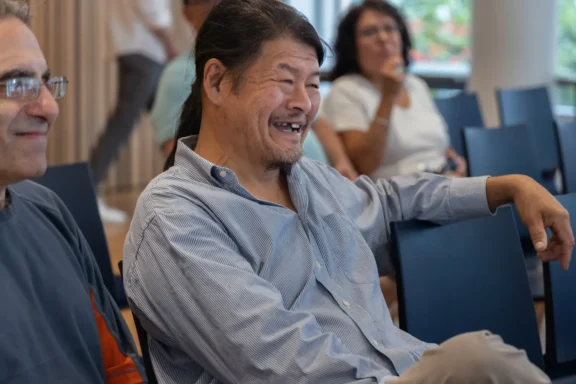 A man sits in a chair smiling and laughing during an indoor event, with other people seated in the background.