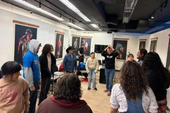 A group of people stands in a circle in a room with portrait artwork on the walls; one person raises their arms above their head.