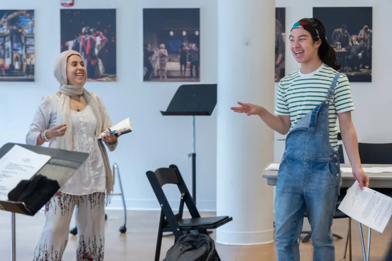 Two people stand indoors, holding scripts and smiling, with theater posters on the wall and music stands and chairs in the background.