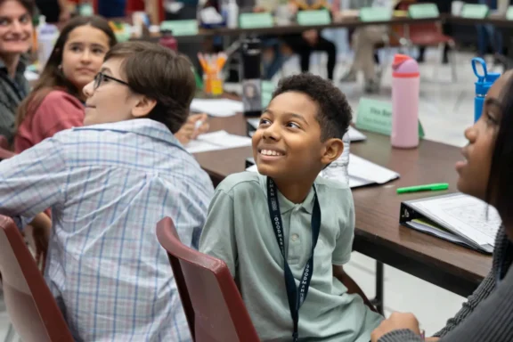 A group of students sit at a classroom table, looking toward the front of the room. Notebooks, water bottles, and school supplies are on the table.