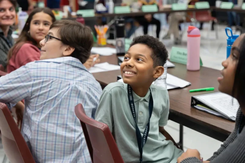A group of students sit at a classroom table, looking toward the front of the room. Notebooks, water bottles, and school supplies are on the table.