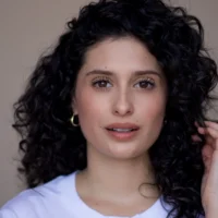 A woman with curly dark hair, wearing a white top and gold hoop earrings, looks at the camera against a plain background.