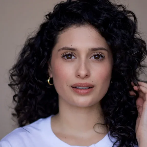A woman with curly dark hair, wearing a white top and gold hoop earrings, looks at the camera against a plain background.