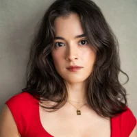 Woman with wavy brown hair wearing a red top and a gold necklace stands against a plain background, looking directly at the camera with a neutral expression.