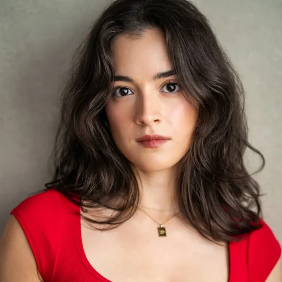 Woman with wavy brown hair wearing a red top and a gold necklace stands against a plain background, looking directly at the camera with a neutral expression.