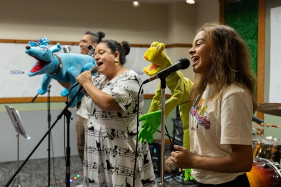 Three people hold colorful puppets and sing into microphones in a room with musical instruments and a whiteboard in the background.