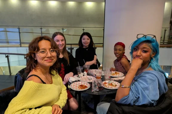 Five people sit around a table with food and drinks, smiling at the camera in a well-lit indoor setting.