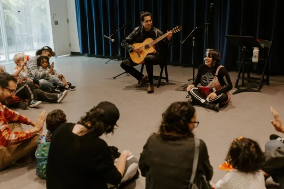 A man playing guitar and a woman sit in front of a group of children and adults in a room with blue curtains, engaging in a musical activity.